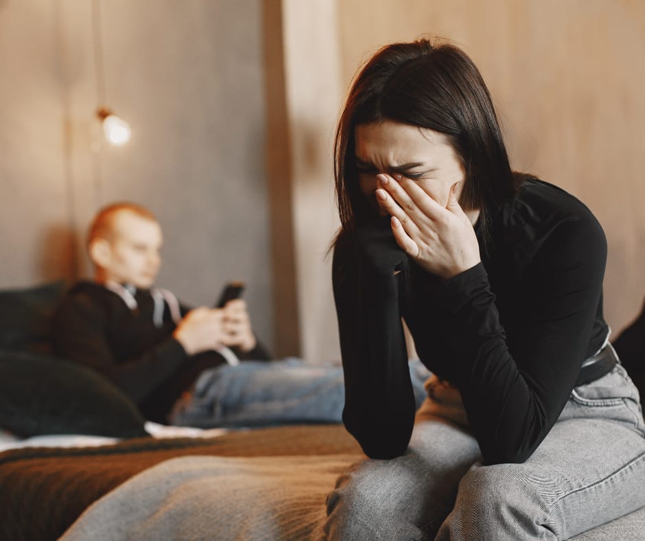 Kuvassa nainen itkee ja pitelee kasvojaan käsillään. Taustalla mies istuu sängyllä puhelin kädessään.