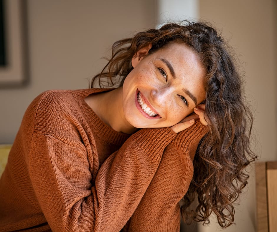 Kuvassa hymyilevä kiharapäinen nainen oranssinruskeassa villapaidassa.