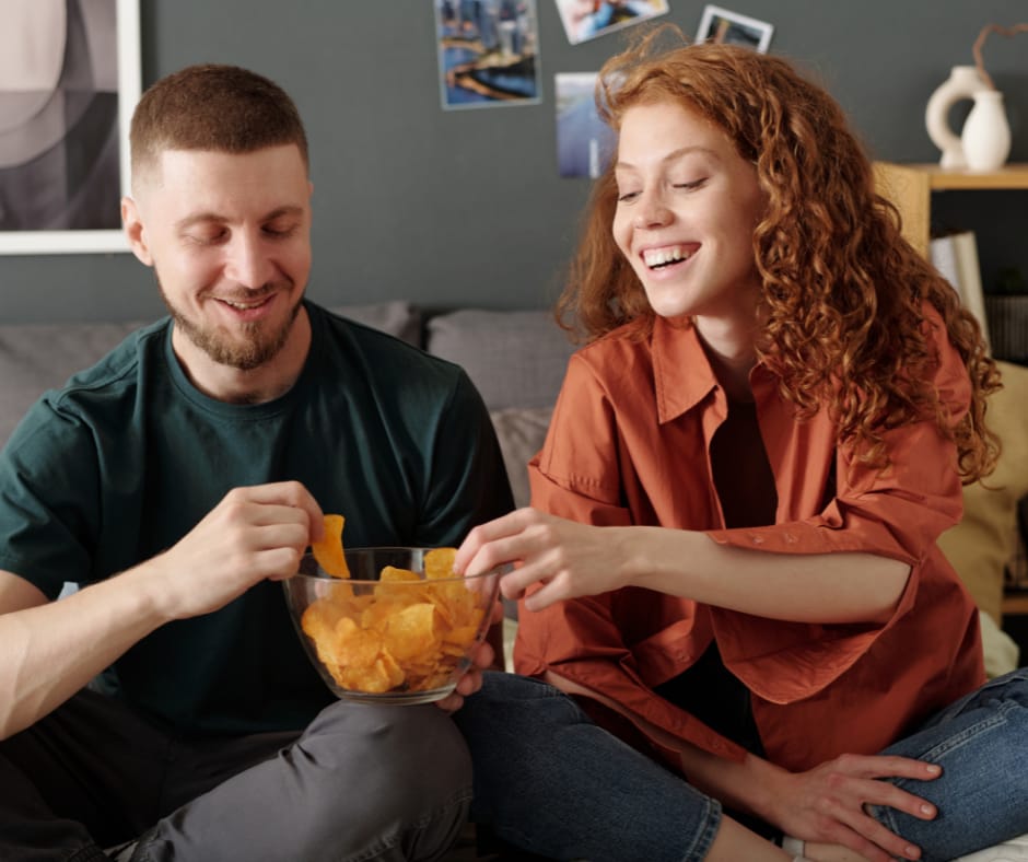 Kuvassa iloinen mies ja nainen syövät sohvalla istuen sipsejä.