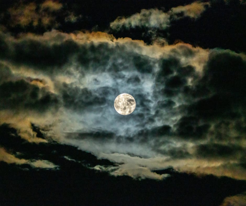 Kuvassa näkyy täysikuu, joka pilkottaa pimeällä taivaalla pilvien takaa.