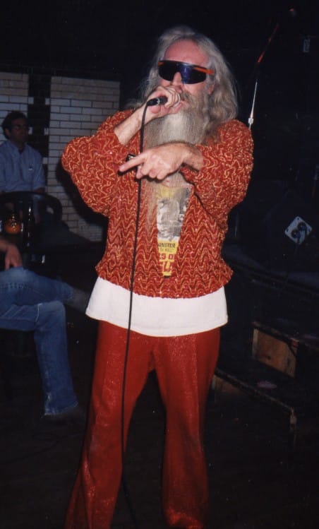 Artisti Johnny Legend esiintyy punaisessa asussa ja aurinkolaseissa. Hänellä on pitkä, harmaa parta. 