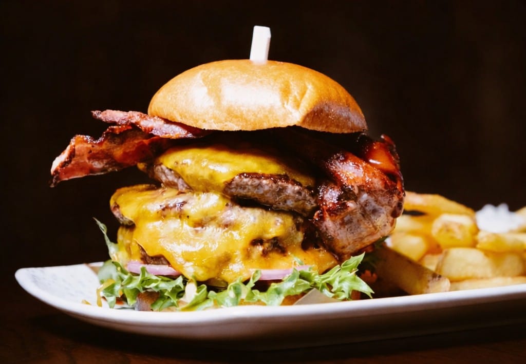 Hampurilainen, jossa on kaksi naudanpihviä, sulatettua juustoa, pekonia ja salaattia, lautasella ranskalaisten kanssa.