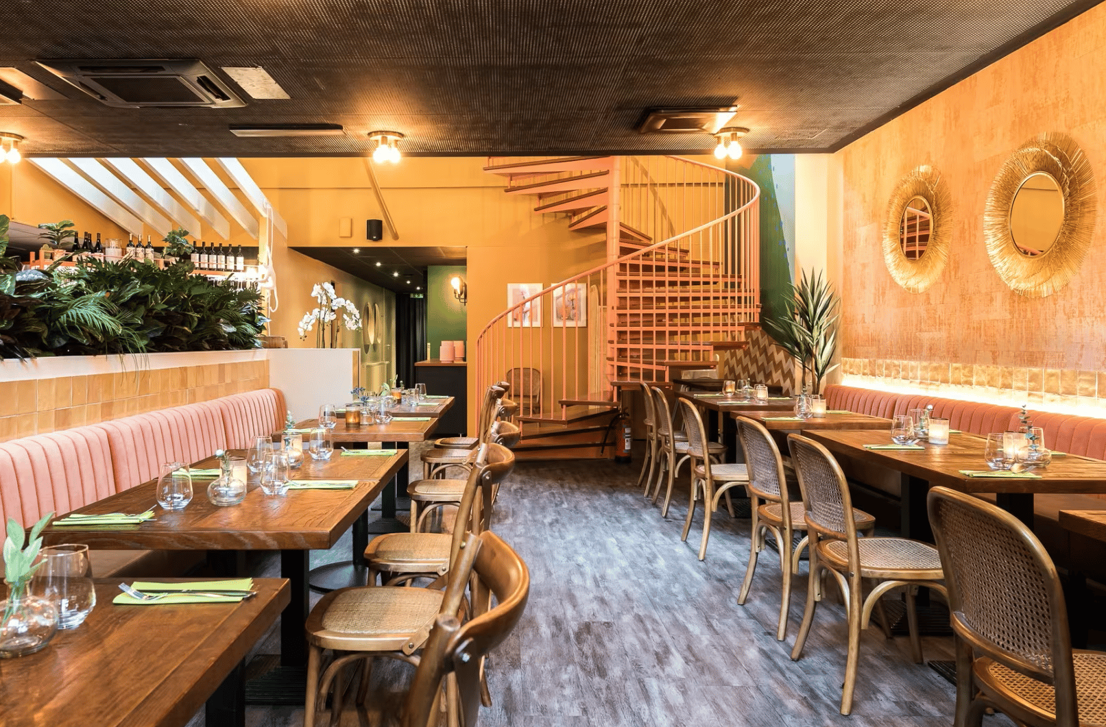 Ravintolan sisätila, jossa on puiset pöydät ja tuolit, vaaleanpunaiset penkit sekä kierreportaat taustalla.