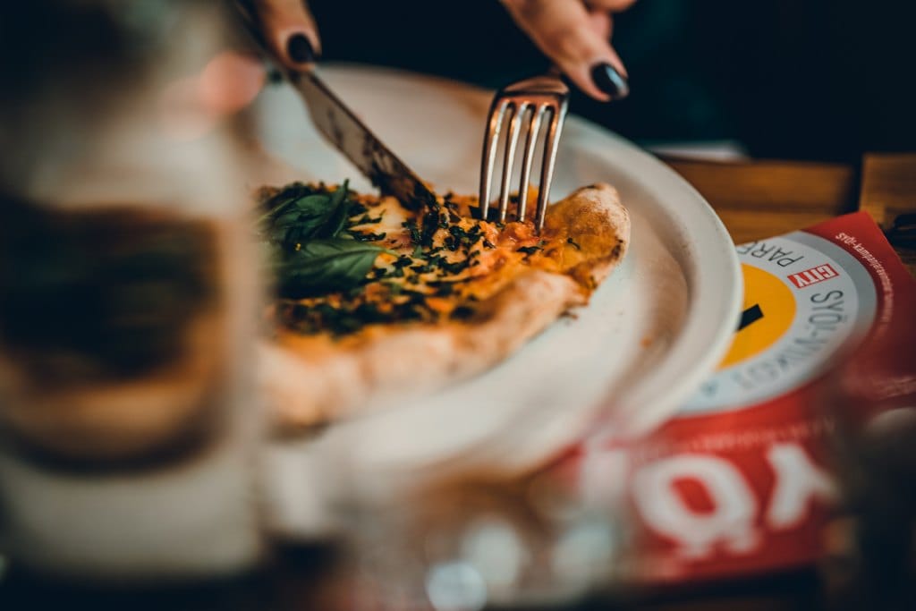Lähikuva henkilöstä leikkaamassa pizzapalaan veitsellä ja haarukalla ravintolapöydässä; etualalla juomalasi ja taustalla näkyy Syö!-viikot-kampanjan materiaalia.