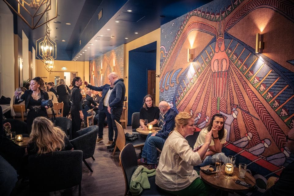 Vilkas ravintolasali, jossa ihmiset istuvat pienissä pöytäryhmissä keskustelemassa ja juomassa. Seinää koristaa suuri, värikäs muraali, ja tunnelma on lämmin ja iltaisa.