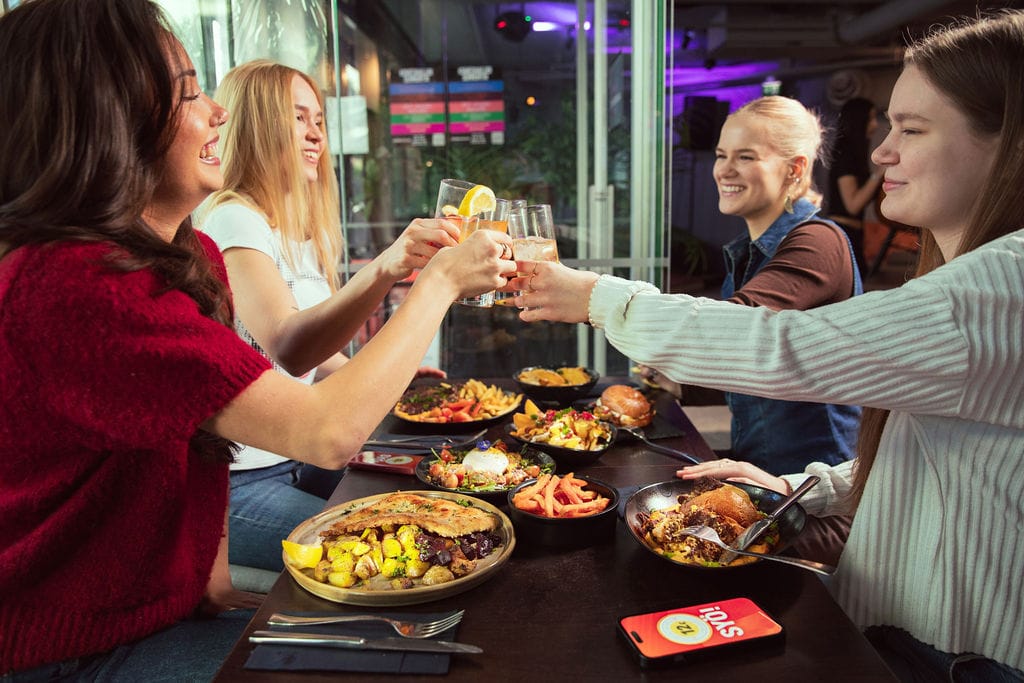Ravintolapöytä täynnä ruokia. Ihmiset kilistävät laseja.