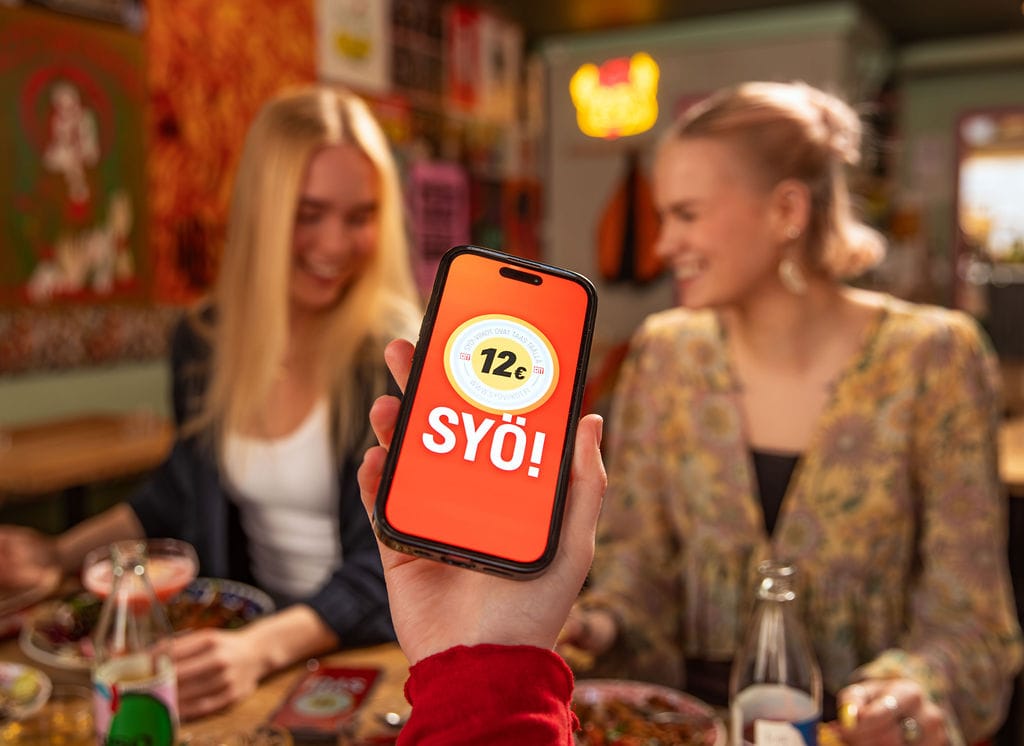 Henkilöitä syömässä ravintolassa. Käsi pitelee puhelinta, jossa näytöllä SYÖ!-viikkojen logo.