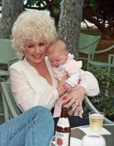 Nuori Dolly Parton istuu ulkona vauva sylissään vaaleassa puserossa ja farkuissa