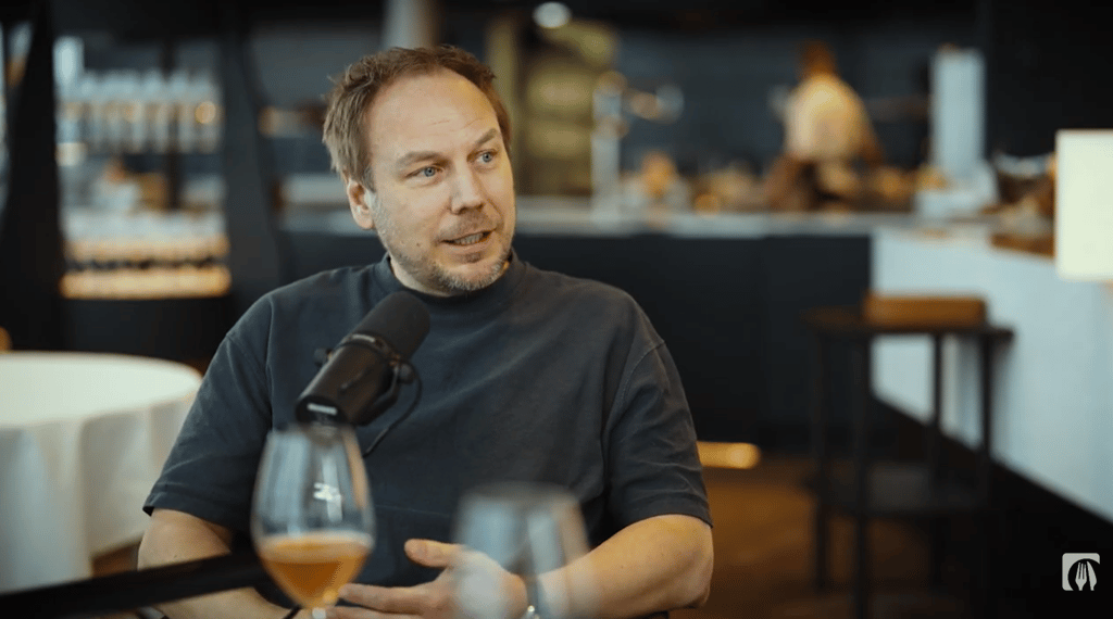 Keittiömestari Tommi Tuominen istuu mikrofoni edessä ravintolasalissa lasi edessä.