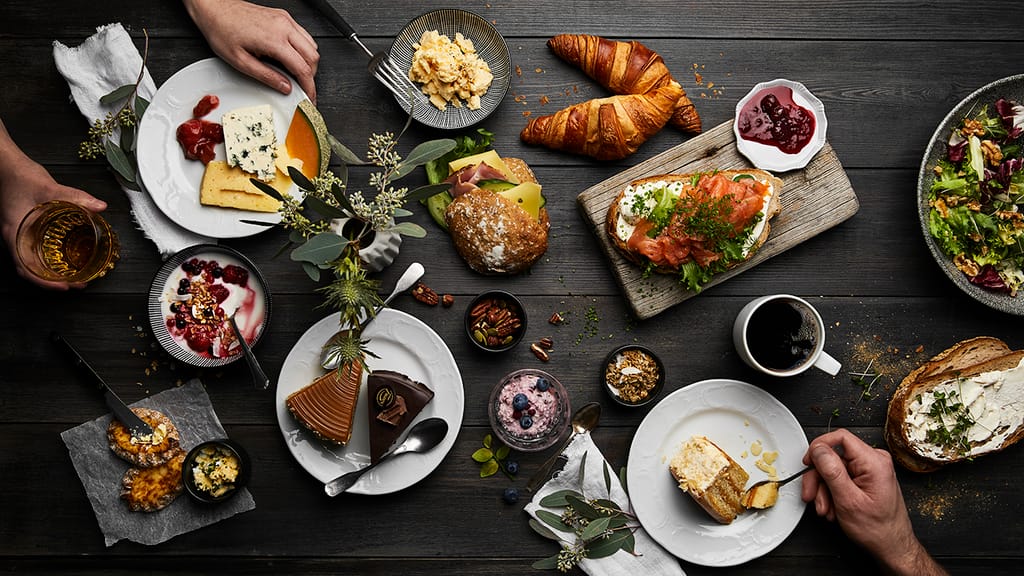 Pöytä täynnä aamiaisherkkuja, kuten croissantteja, leipiä ja kakkuja.