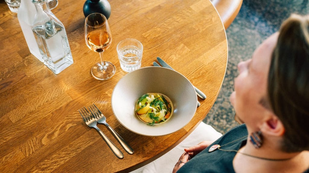 Nainen istuu ravintolapöydässä, edessä annos, aterimet ja vettä.