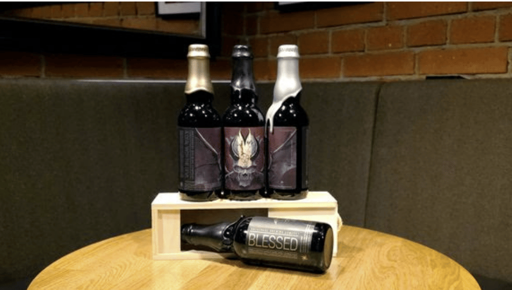 Kolme tummaa olutpulloa esillä puisella telineellä ja yksi pullo kyljellään pöydällä, taustalla tiiliseinä ja pehmustettu penkki ravintolassa.