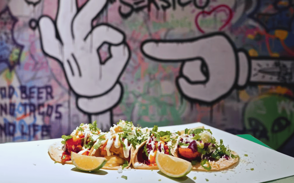 Kolme täytettyä tacoa lautasella, päällä yrttejä ja kastiketta, taustalla käsiaiheinen graffitimaalaus.