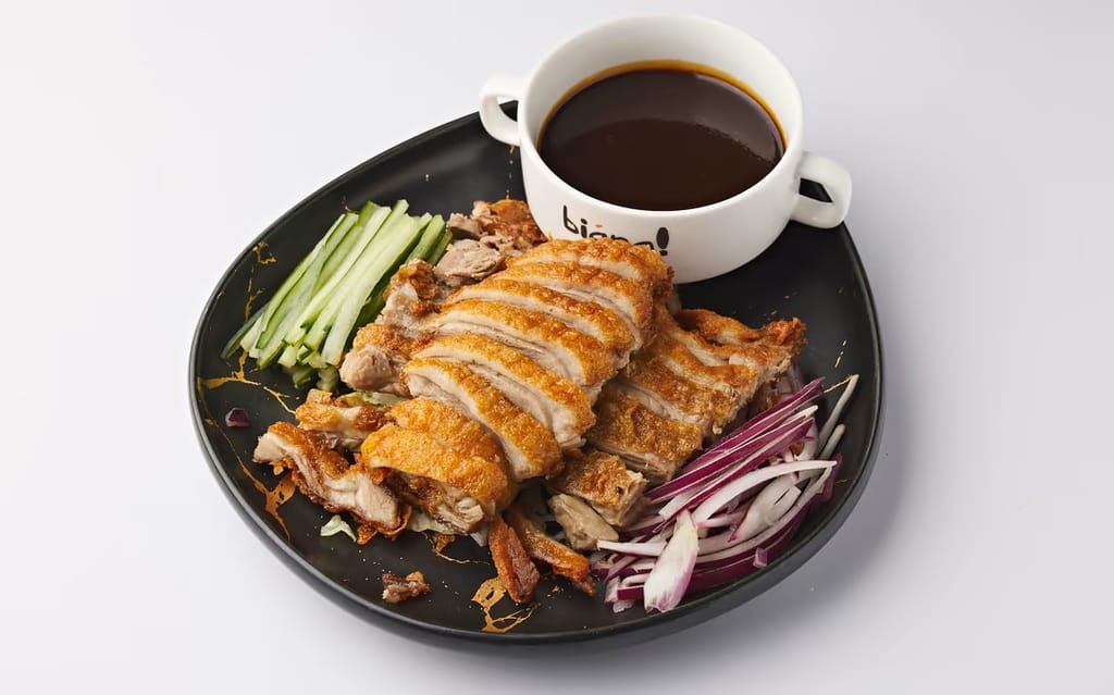 Viipaloitua rapeakuorista ankkaa lautasella, vierellä kurkkusuikaleita, punasipulia ja kulhollinen tummaa kastiketta.