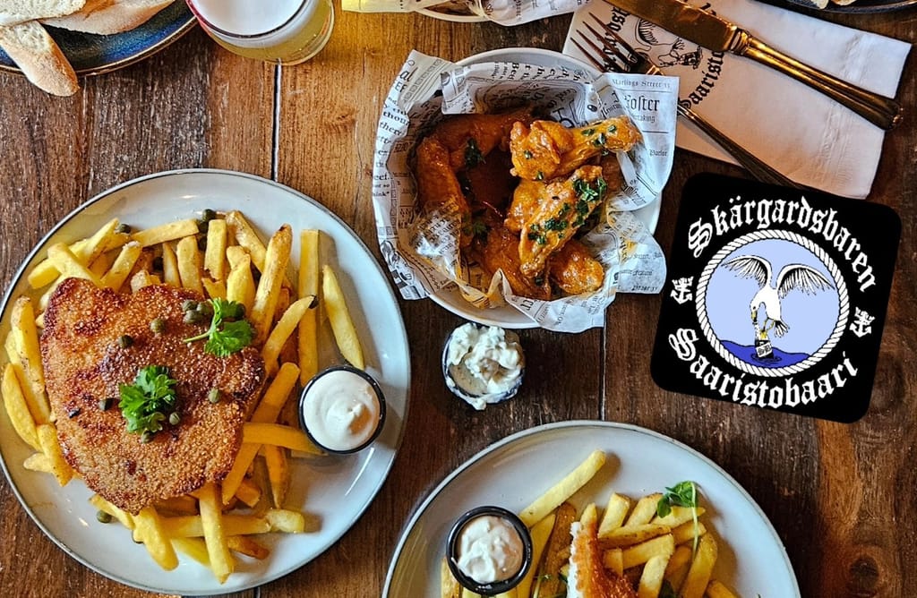 Pöydällä annoksia: leivitetty schnitzel ranskalaisten ja kastikkeen kanssa, kulhollinen kanansiipiä sekä dippejä.