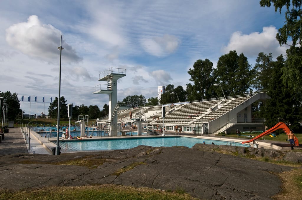 Helsingin Uimastadionin hyppytorni ja lastenallas vesiliukumäkineen, etualalla kallio ja taustalla katsomo ja siniset liput.
