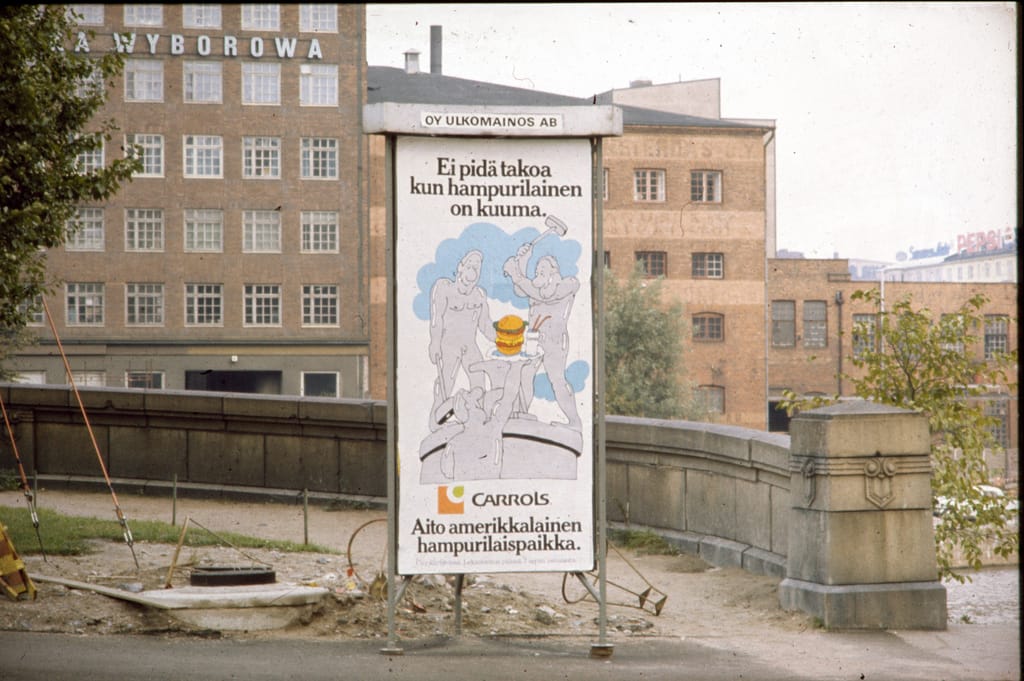 Vanha ulkomainostaulu kaupunkimaisemassa. Mainoksessa on Carrols-hampurilaisravintolan piirroskuva ja teksti ”Ei pidä takoa kun hampurilainen on kuuma.” Taustalla näkyy tiilirakennuksia ja teollinen ympäristö, mikä viittaa menneeseen vuosikymmeneen.