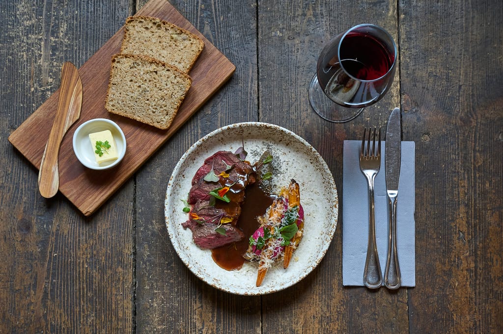 Lautasella liha-annos kastikkeella ja paahdetuilla kasviksilla, vieressä leipää, voita ja lasi punaviiniä rustiikkisella pöydällä.