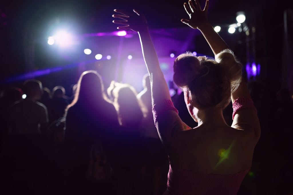 K-Pop-henkisesti pukeutunut nainen seisoo värivalojen loisteessa keikalla ja on kädet pystyssä. Naisella on värikäs paita ja blondattu tukka joka on kahdella nutturalla pään päällä. 