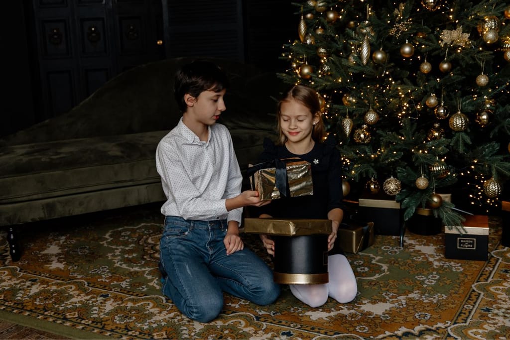 Nostalginen, ysärimäistä tunnelmaa jäljittelevä kuva, jossa lapset istuvat joulukuusen juurella jakamassa paketteja. Tyttö ja poika. 