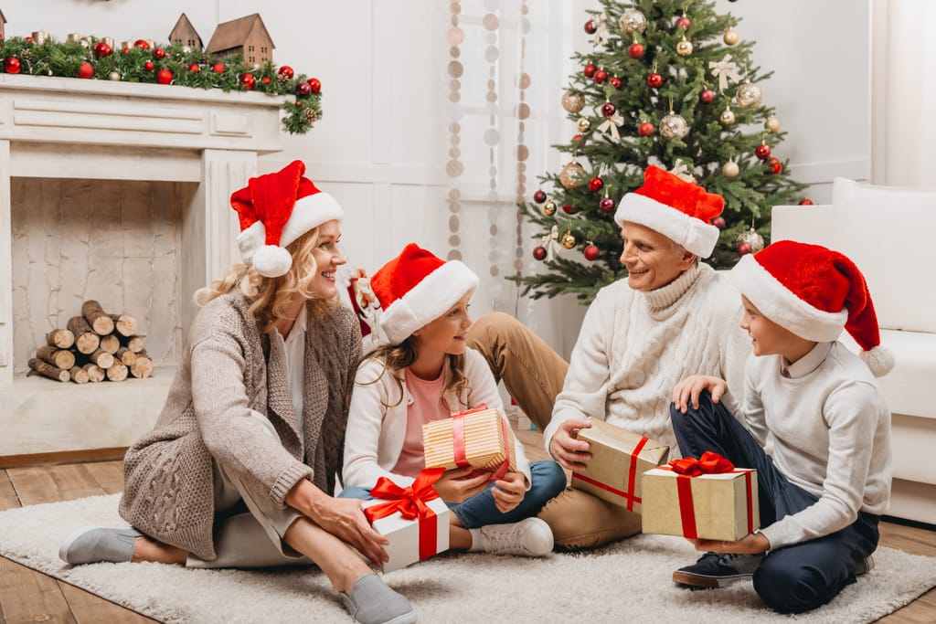 Perhe viettämässä joulua valkoisessa ja minimalistisessa kodissa. Kaksi lasta, äiti ja ilmeisesti isoisä samassa kuvassa. kaikilla karvareunuksinen tonttulakki päässä ja jokaisella perinteisesti paketoitu lahja kädessä. Taustalla oleva takka on tekotakka muutamalla klapilla ja taustalla näkyy joulukuusi. Valkoinen sohva. Kuvituskuva jouluteemaisessa artikkelissa. 