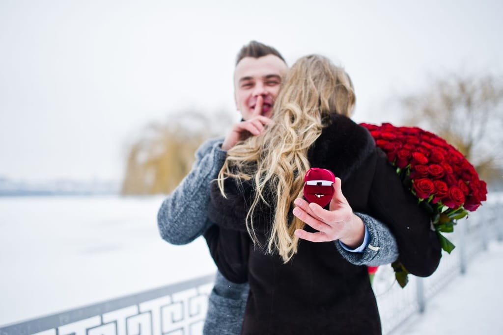 Mies kosii tyttöystäväänsä ja näyttää sormin hys iso kukkapuska kainalossa. Halaus ja sormusrasia. 