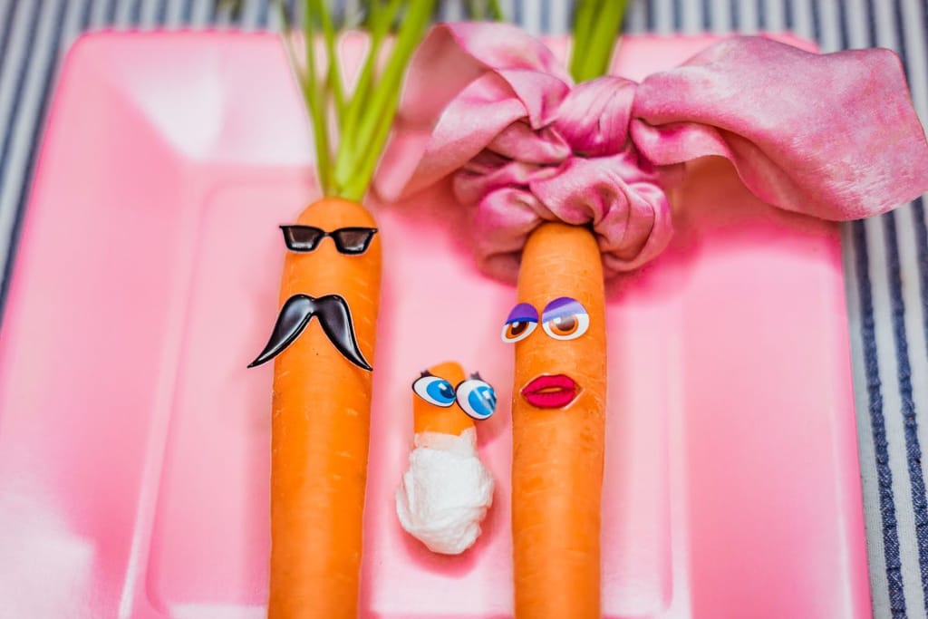 Herra ja rouva porkkana ovat saaneet pienen vaippoihin laitetun porkkanan, joka tuijottaa äitiporkkanaa. Kuvituskuva. Kuvassa on kaksi porkkanaa pinkillä lautasella, ja toiselle on laitettu tarrasta viikset ja toiselle silmär ja silmät ja suu tarroista. Vauvaporkkanalla on olevinaan vaipat ja silmät. 