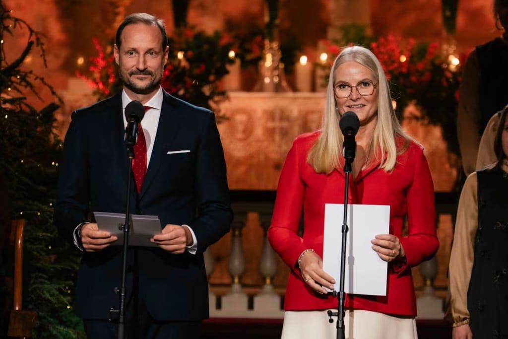 Norjan kruununprinssipari pitämässä puhetta virallisessa joulutilaisuudessa, jossa kiitettiin pelastusviranomaisia. Kuvassa Mette-Marit silmälasit päässä, pitelee puhetta kädessään ja puhuu mikrofoniin. Vaaleat hiukset. Mies kaljuuntuva, puku päällä. Molemmilla mikrofonit edessään. Tausta punainen ja jouluinen, tunnelmallinen valaistus. 