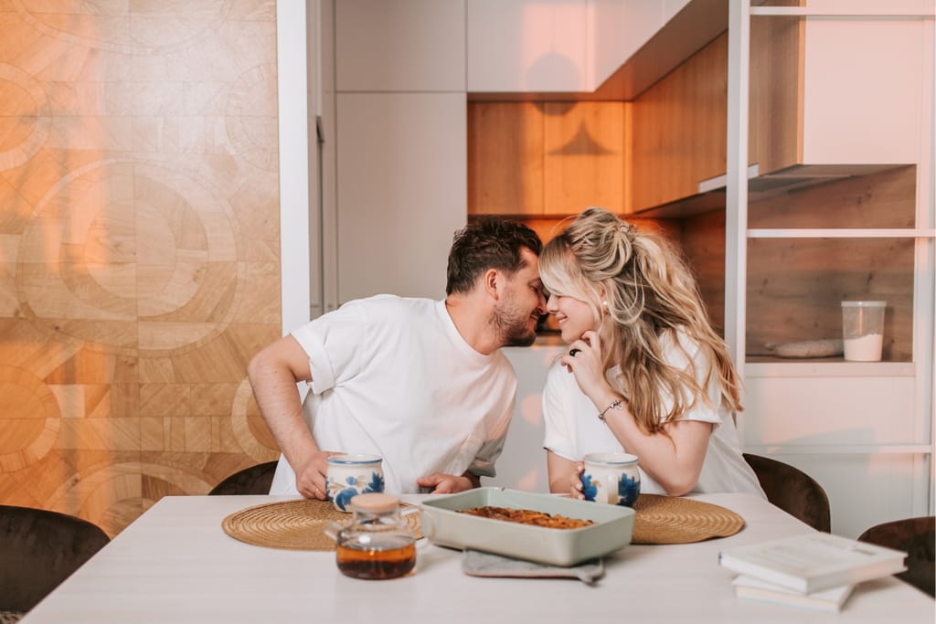 Pariskunta istuu keittiön pöydän ääressä ja nojautuu toisiaan kohti, otsat vastakkain ja hymy huulilla. He pitävät kahvimukeja käsissään, ja pöydällä on teekannu sekä uunivuoka. Kuvassa välittyy kodikas ja rakastava hetki arjen keskellä.