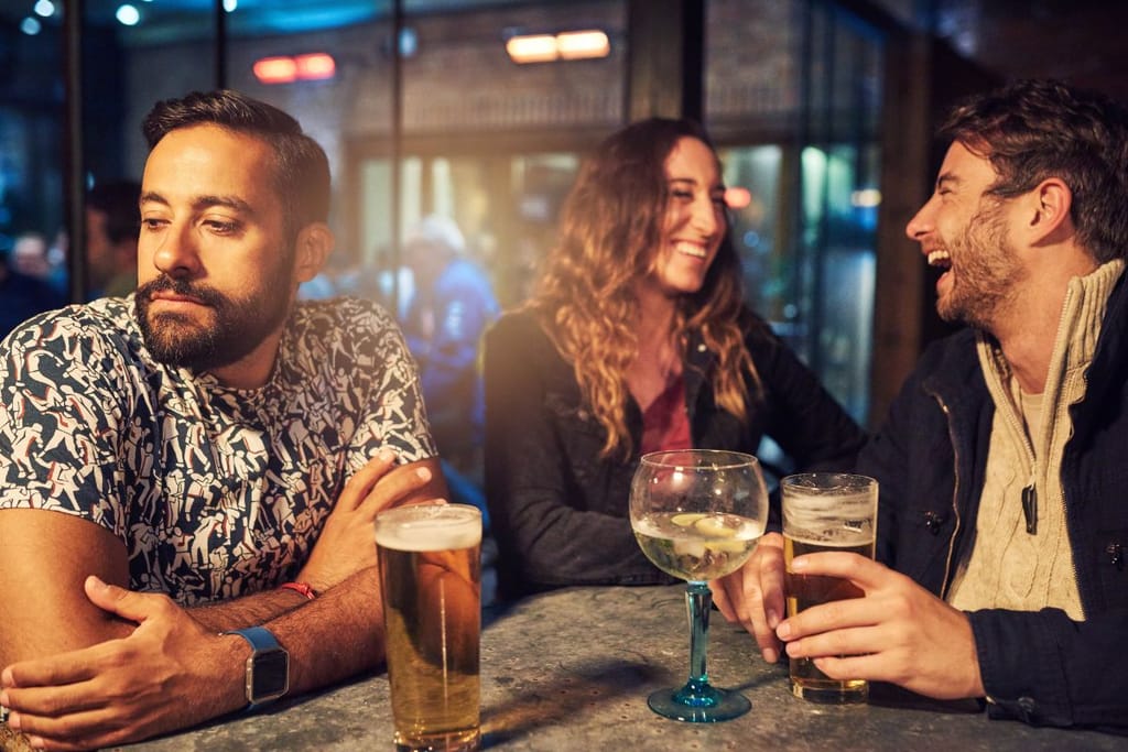 Pariskunta istuu baarissa kolmestaan ja yksi on ulkopuolinen. Mies ja nainen juttelevat ja nauravat keskenään ja mies istuu poispäin muista. 