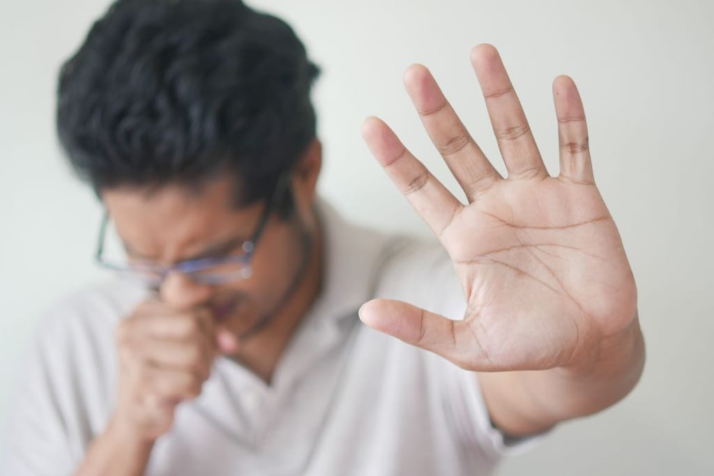 Mies yskii käteensä ja näyttää ei-käsimerkkiä kuvassa. Kuvituskuva yskää koskevalle artikkelille