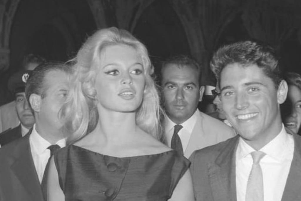Brigitte Bardot tapahtumassa ihmisten keskellä vastanäyttelijänsä kanssa. Hän on mustavalkoisessa kuvassa nuorena hiukset auki, ei katso kameraan ja toinen etualalla vasemmalla kuvaajasta katsoen. 