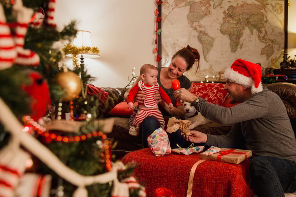 Kuvituskuva perheestä, joka viettää joulua ja jakaa lahjoja toisilleen. Kuvassa on joulukoristeltu kuusi ja isällä tonttulakki ja vauva on ihmeissään uudesta lelusta.  Öiti pitää sylissä. 