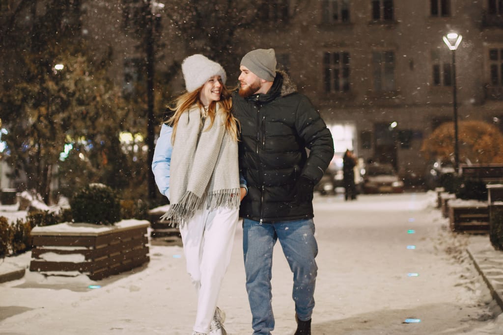Talvinen iltakatu, jossa nuori pari kävelee vierekkäin käsikynkässä. Molemmilla on talvivaatteet, pipot ja huivit, ja he hymyilevät toisilleen lumisateessa valaistulla kaupunkipolulla.