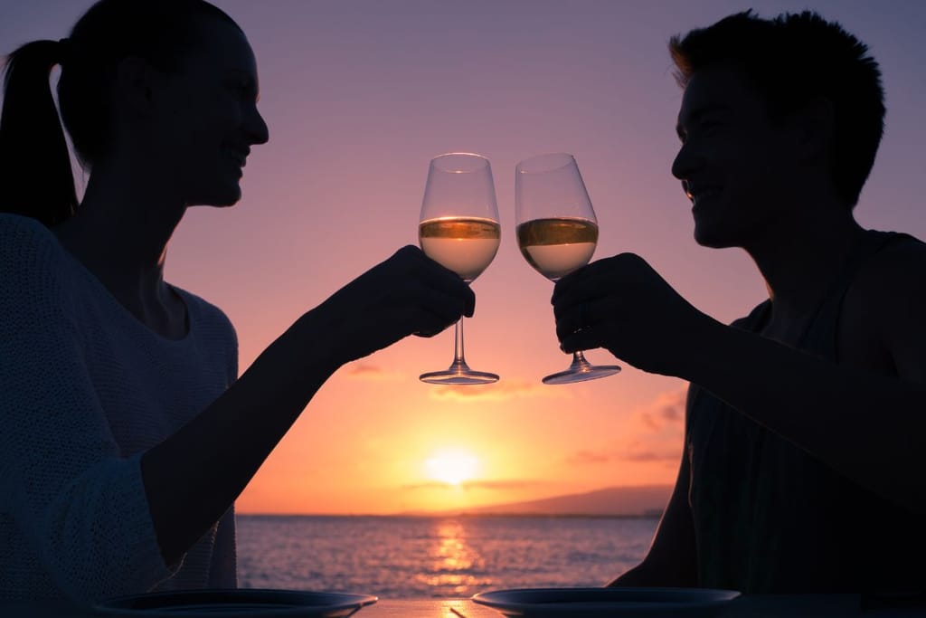 Nainen ja mies juo auringon laskussa viiniä ja kilistelevät. heistä näkyvät vain mustat siluetit loppuillan aurinkoista merenselkää vasten. 