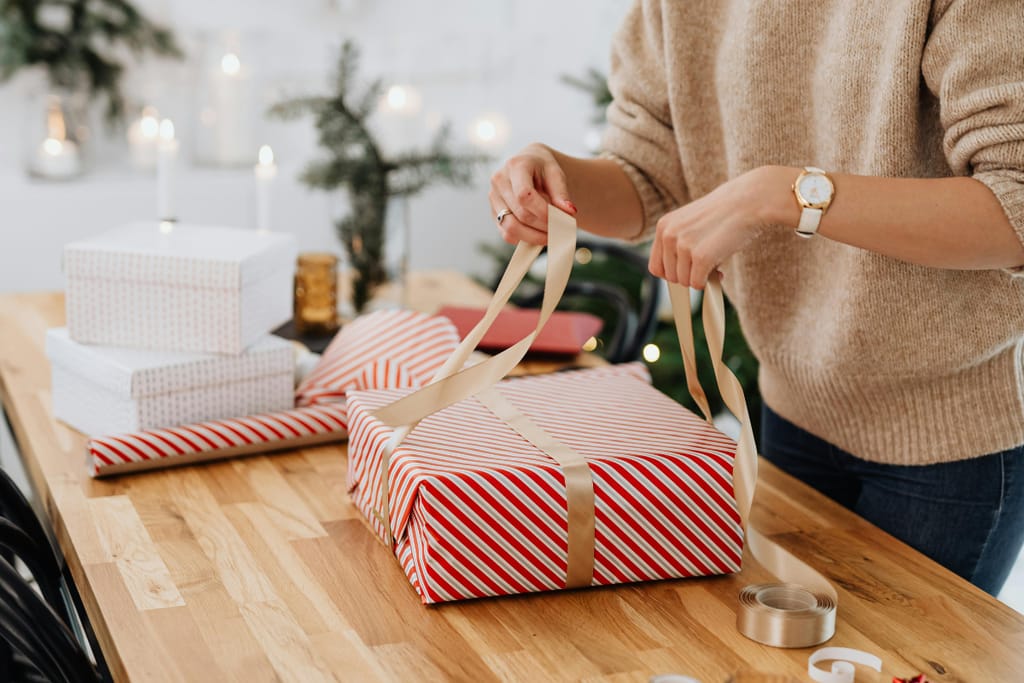 Nainen paketoi joululahjaa silkkinauhalla. Joulupaketti on perinteisellä paperilla päällystetty paketti ja naisella on ranteessaan valkokullanvärinen rannekello, vaatteena beigen värinen neule ja tummat farkut. Pöytä on koivupuuta. Sopisi Ikean kuvastoon tai mainokseen, mutta menee nyt jouluteemaisessa artikkelissa kuvituskuvana. 