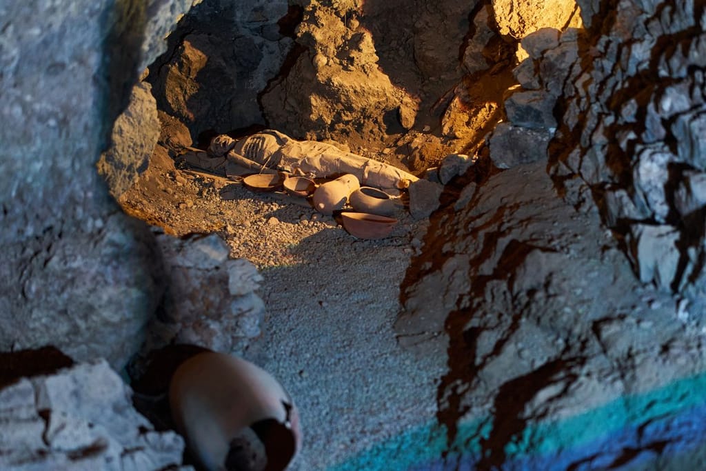 Kuvassa luola, jossa jonkun jäänteet ja astioita. Kuva on hämärä ja otettu pienessä valossa. Muumiolla on yllään peite. 