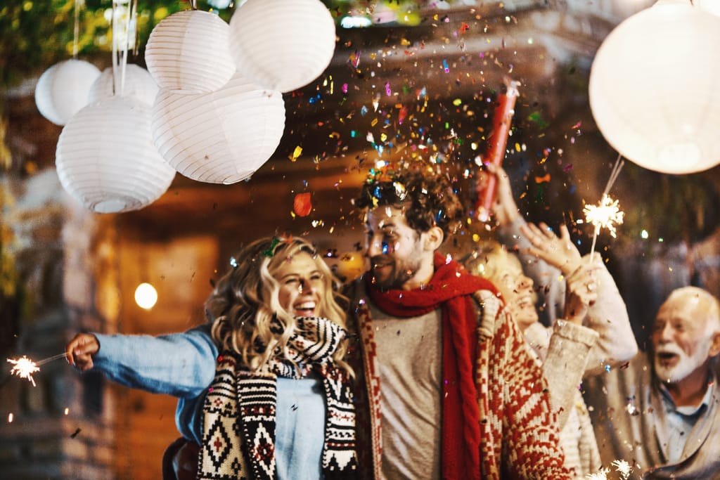 Ryhmä ihmisiä juhlii ulkona illalla paperilyhtyjen alla, kädessä tähtisädetikkuja, ilmassa leijailee konfettia ja ihmiset nauravat ja liikkuvat yhdessä.