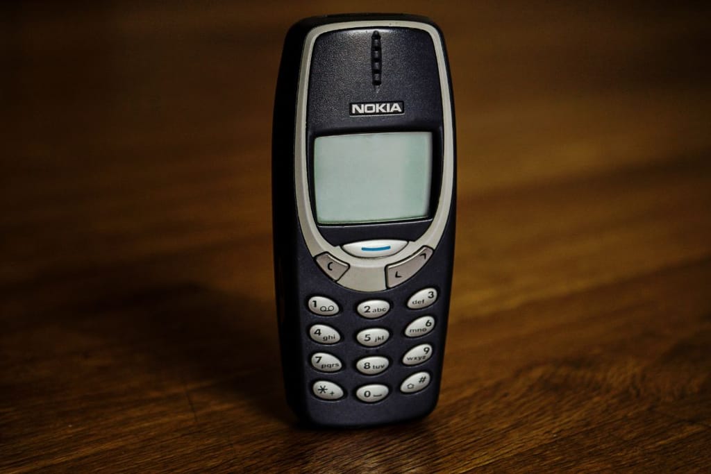Vanha nokialainen eli nokian puhelin, joka kesti kylmää, märkää eikä puhelin palanut helvetin tulessakaan ja puhelin oli simppeli käyttää, mutta akku oli kestävä. Puhelin on ruskealla pöydällä, hopeanvärinen näppäimistö ja sammunut ruutu. Jokainen muistaa näiden soittoäänen.