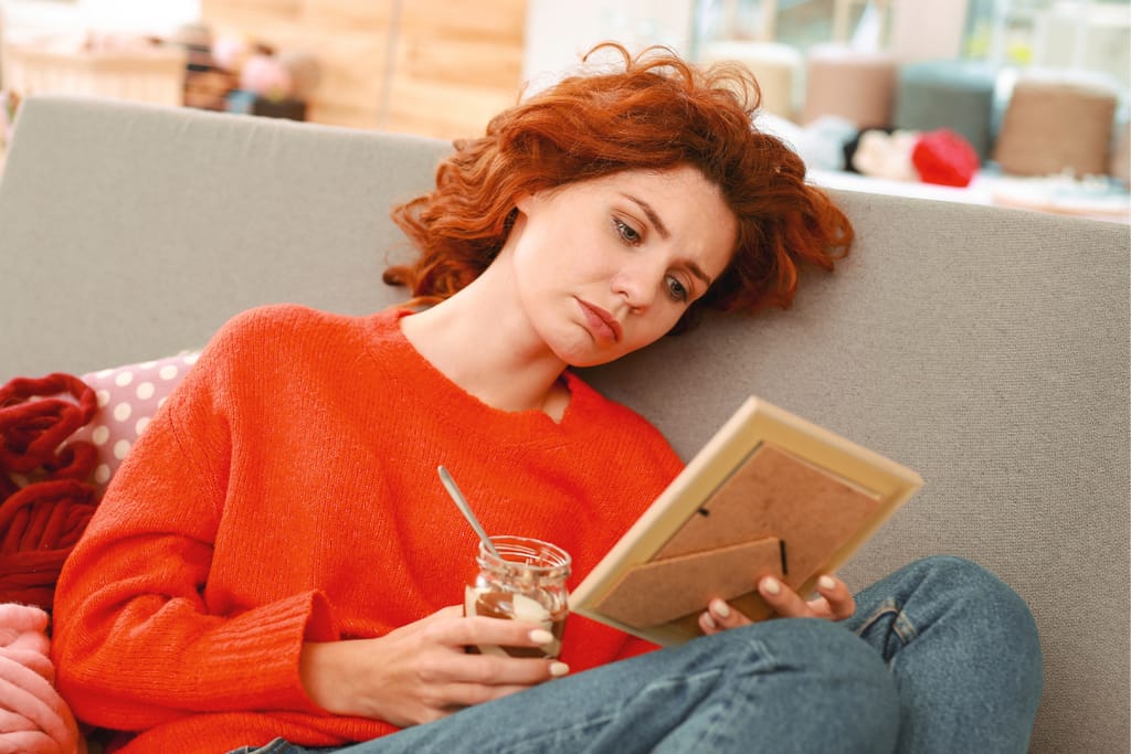 Kuvassa punahiuksinen nainen istuu sohvalla ja katselee kaihoisasti kuvaa exästää. Hänellä on toisessa kädessään juomalasi.