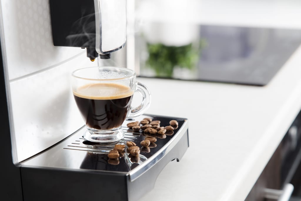 Espressokeitin kapselikeitin kotona pöydälllä. Kuppiin valuu mustaa kahvia ja pöydällä on myös kokonaisia kahvipapuja.