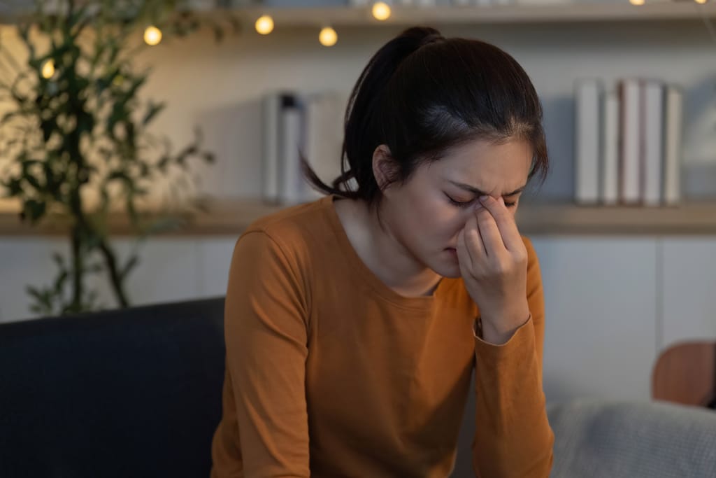 Nuori nainen istuu sisätiloissa ja hieroo silmiään näyttäen väsyneeltä tai stressaantuneelta.