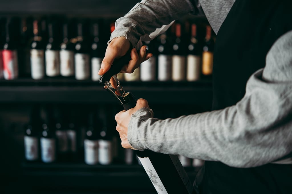 Kuvassa baarimikko avaa viinipulloa. Taustalla näkyy viinipulloja hyllyssä.
