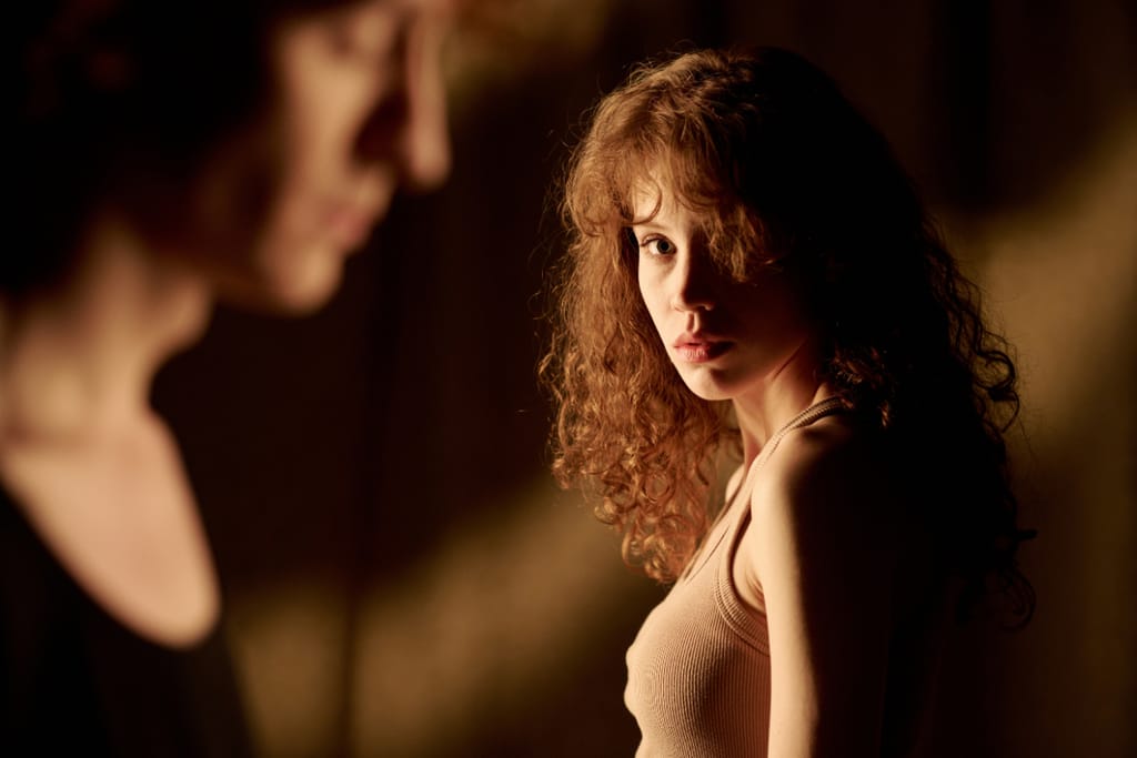 Nainen seisoo hämärässä valaistuksessa ja katsoo sivulle päin, etualalla näkyy toisen henkilön kasvot epätarkkoina, tausta on tumma ja lämminsävyinen.