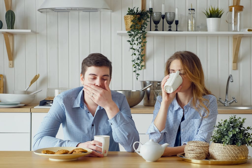 Pariskunta istuu aamiaisella keittiön pöydän ääressä väsyneinä ja etäisinä. Mies haukottelee ja peittää suunsa kädellään, nainen juo kahvia katse poispäin. Pöydällä on teekannu, kuppeja ja lautasella keksejä. Ilmapiiri on arkinen ja hieman uupunut, kuin keskustelu olisi loppunut ennen kuin se ehti alkaa.