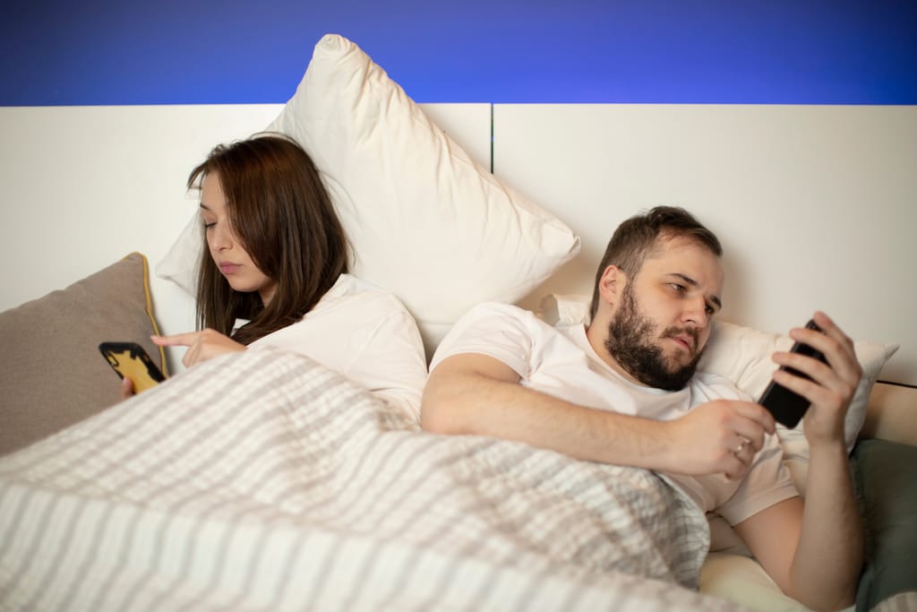 Pariskunta makaa sängyssä selät vastakkain, kumpikin katsoo omaa puhelintaan. Ilmeet ovat väsyneet ja etäiset, eivätkä he ole vuorovaikutuksessa keskenään. Kuvassa korostuu tunne etääntymisestä ja arjen rutiinien hiljaisesta välimatkasta.