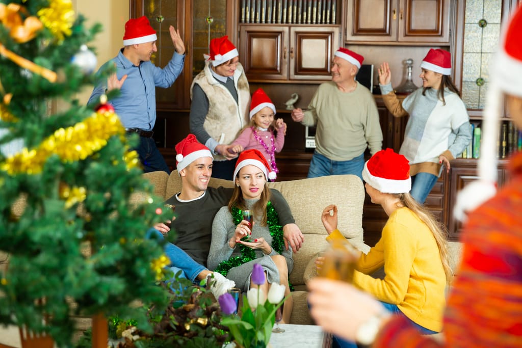 Iloinen perhe ja ystävät viettävät joulua yhdessä olohuoneessa, useimmat heistä käyttävät punaisia joululakkeja. Osa istuu sohvalla juomia käsissään, kun taas taustalla muut tanssivat ja juttelevat. Etualalla näkyy koristeltu joulukuusi ja pöydällä kukkia. Tunnelma on lämmin ja vilkas, täynnä juhlan tuntua.