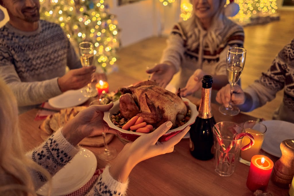 Jouluillallinen kynttilänvalossa: neljä ihmistä istuu pöydän ääressä ja kohottaa maljoja, kun yksi heistä ojentaa vadilla paahdettua kanaa ja kasviksia. Pöydällä on samppanjapullo, juomalaseja ja joulukoristeita. Taustalla näkyy valaistu joulukuusi ja pehmeä, lämmin valaistus luo kodikkaan tunnelman.
