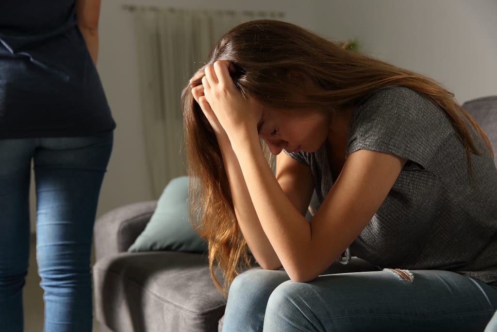Nainen istuu sohvalla kumartunutena pää käsiensä varassa, selvästi ahdistuneena, kun toinen henkilö seisoo taustalla selin ja poistuu huoneesta.