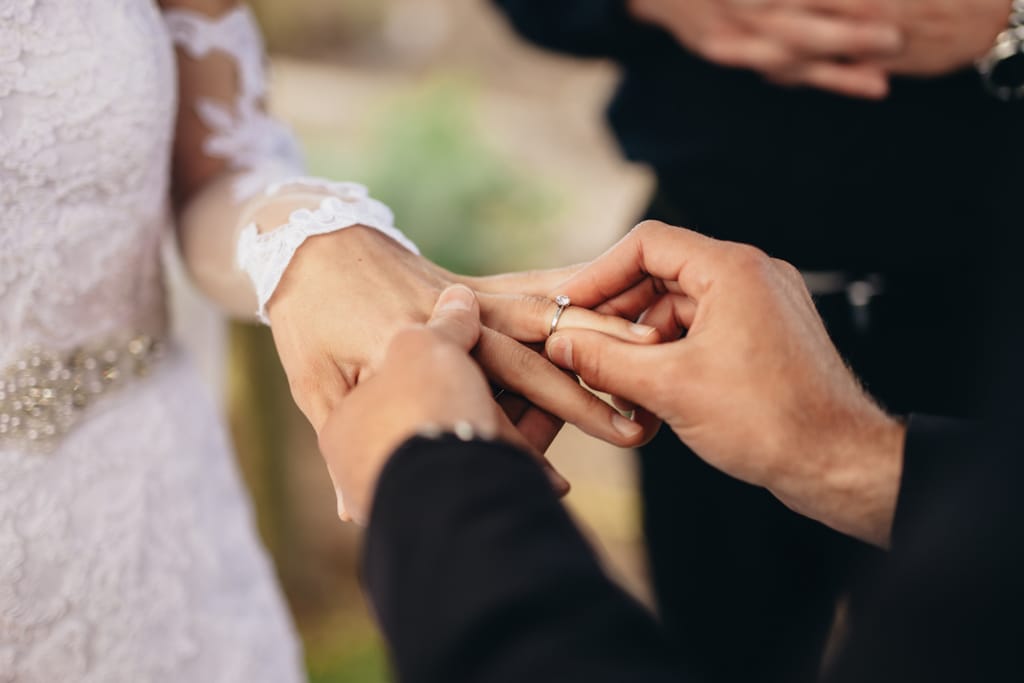 Lähikuva hetkestä, jossa henkilö pujottaa sormuksen toisen sormeen hääseremoniassa. Morsiamella on valkoinen pitsinen häämekko, ja taustalla näkyy hämärästi vihkipaikka. Kuvassa symboloidaan avioliittoa, sitoutumista ja lupauksia.